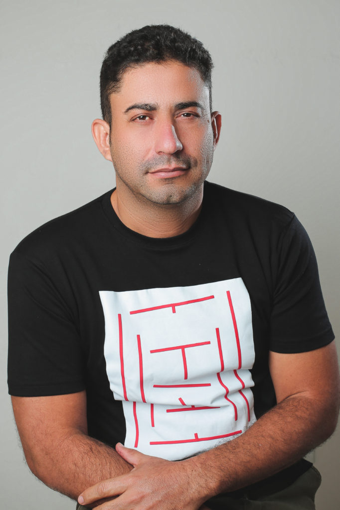 Fabio Alan Locutor olhando fixamente para frente, vestido uma camisa preta com o centro branco e listras no meio vermelhas(parecendo um labirinto), com um fundo de parede com uma cor clara.